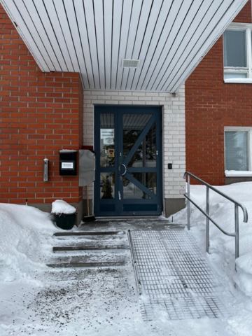 Sosiaalitoimen sisäänkäynnin tummansininen ovi erottuu valkoisesta tiiliseinästä. Muuten rakennus on punatiilinen. Ovelle johtaa suora, jyrkkä luiska ja luiskan vasemmalla puolella portaat, joissa ei ole käsijohdetta.