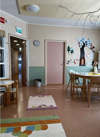 Valoisa leikkihuone, jossa on pöytä, matto ja lasten piirrustuksia seinällä.