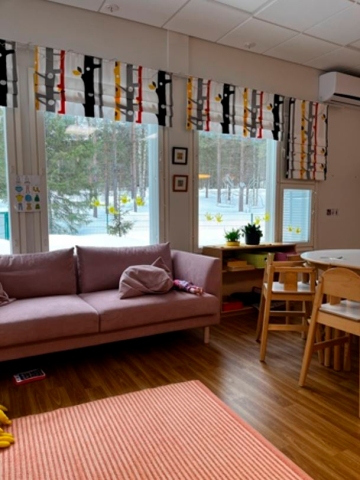 Huoneessa on violetti sohva ja sohvan takana ikkunoissa värikkäät verhot. Lattialla on oranssi matto.