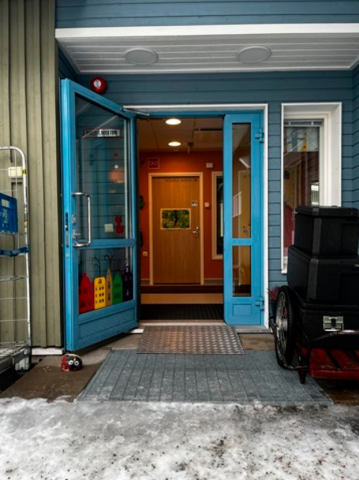 Pääsisäänkäynnin sininen ovi on auki. Metallinen lyhyt ja kapea luiska johtaa ovelle, mutta oven edessä ei ole tasannetta luiskan jälkeen.