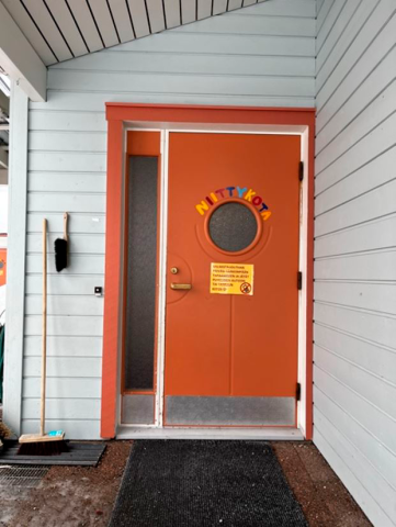 Punainen ovi vaaleaa seinää vasten. Sisäänkäynti on katoksessa ja oven edessä on kuramatto.