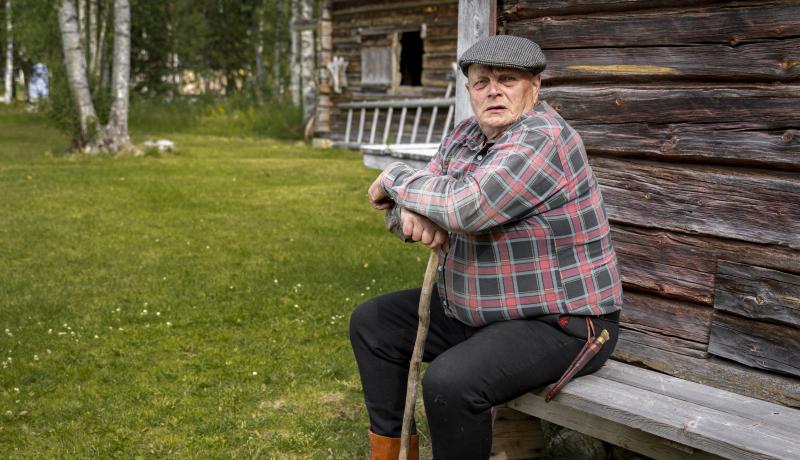 Syytinkiläinen istuskelemassa penkillä keppiin nojaillen