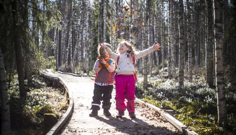 Lapset hymyilee pitkospuilla polulla metsässä.
